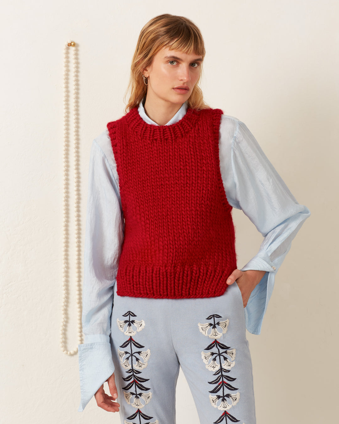 A person wears the Quentin Cardinal Knit Top, a luxurious hand-knitted vest made of merino wool, over a light blue shirt with wide cuffs and patterned trousers, standing near a hanging pearl necklace.