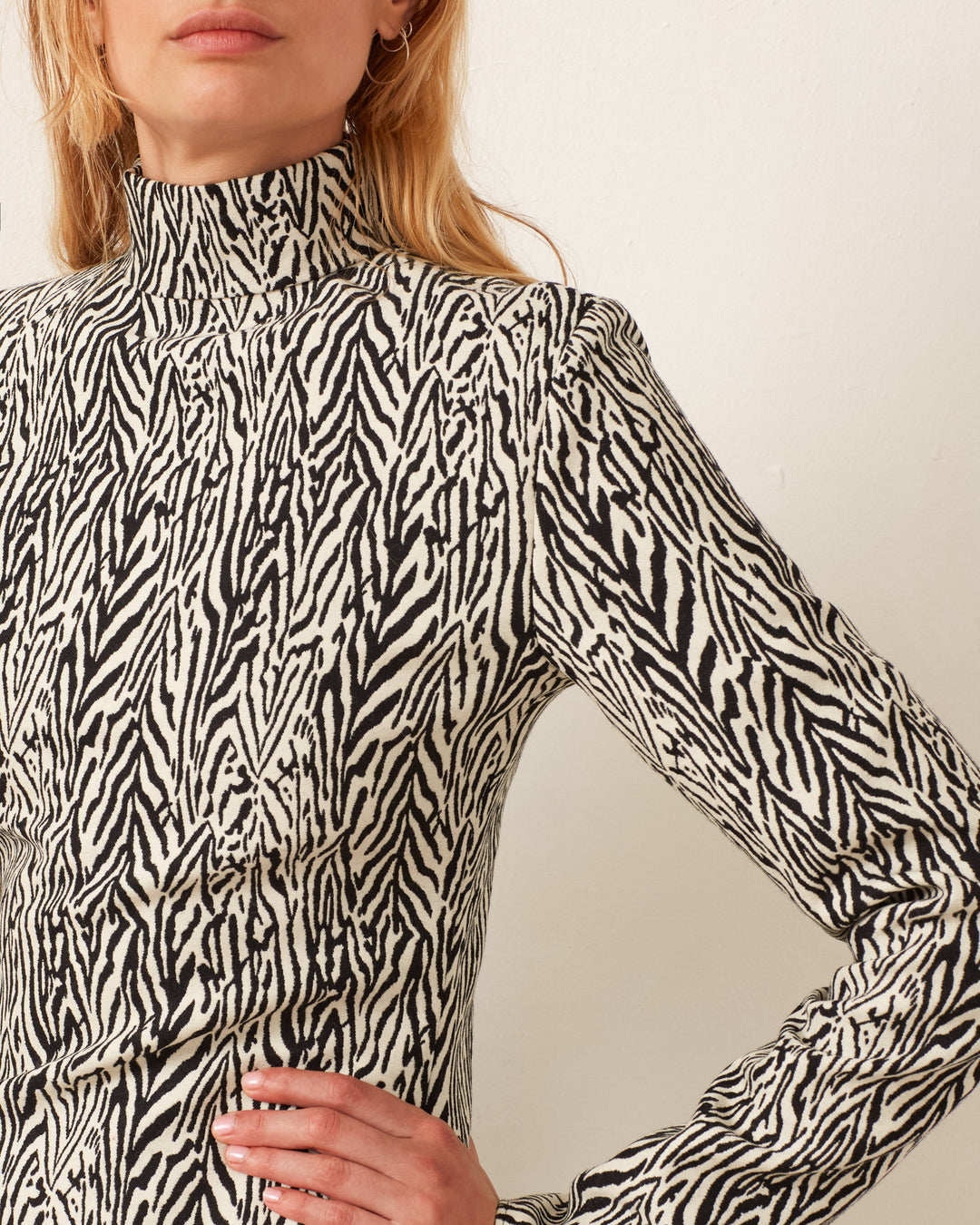 A person is dressed in the Romy Zebra Dress, featuring a high-neck and long sleeves. It is made from a luxurious blend of virgin wool and silk, showcasing a striking black and white zebra stripe pattern.