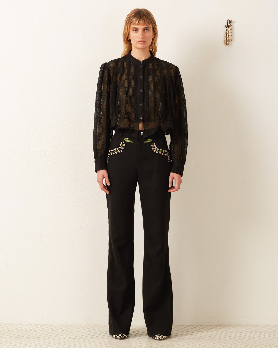 A person stands against a plain white background wearing the Ryland Blackjack Daisy Shirt, a black sheer button-up blouse with floral lace, paired with high-waisted black pants featuring intricate embroidery. Their long hair cascades as they gaze directly at the camera, exuding a modern twist on the Victorian silhouette.