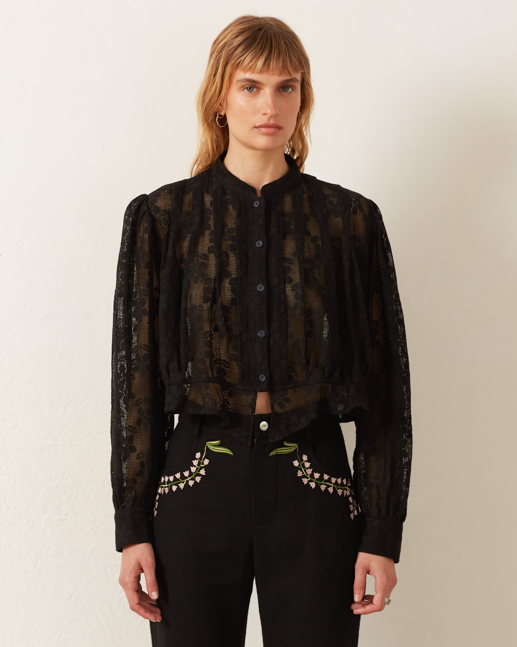 A person with shoulder-length hair wearing the Ryland Blackjack Daisy Shirt stands against a plain white background. The sheer black fabric flows gracefully, paired with black pants adorned with floral embroidery on the pockets, adding an elegant touch to the ensemble.