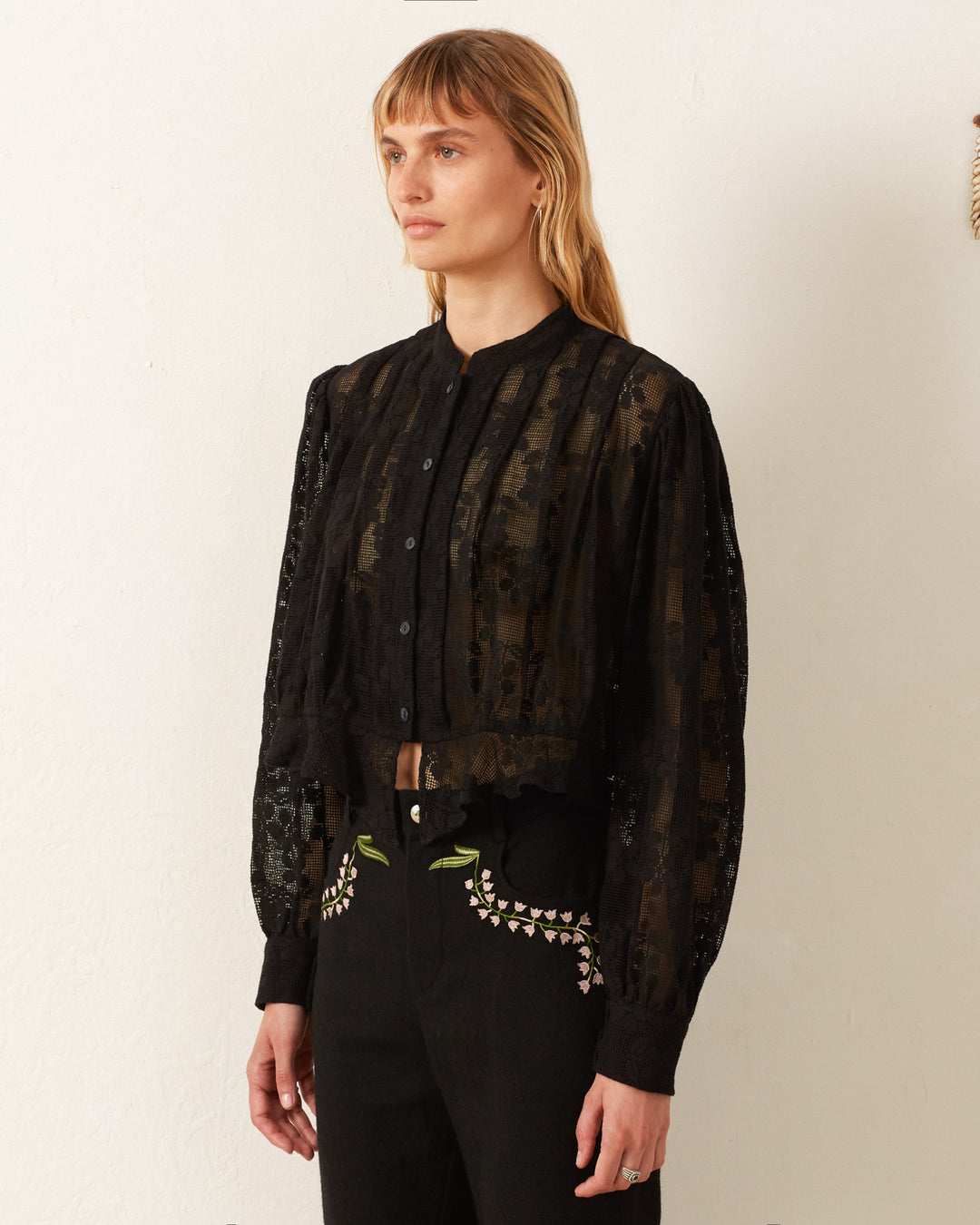 A person dons the Ryland Blackjack Daisy Shirt, a black, semi-sheer blouse with a stand collar and intricate embroidered detailing that evokes a Victorian silhouette. They complement it with black pants adorned with floral embroidery on the pockets, all set against a plain, light-colored wall.