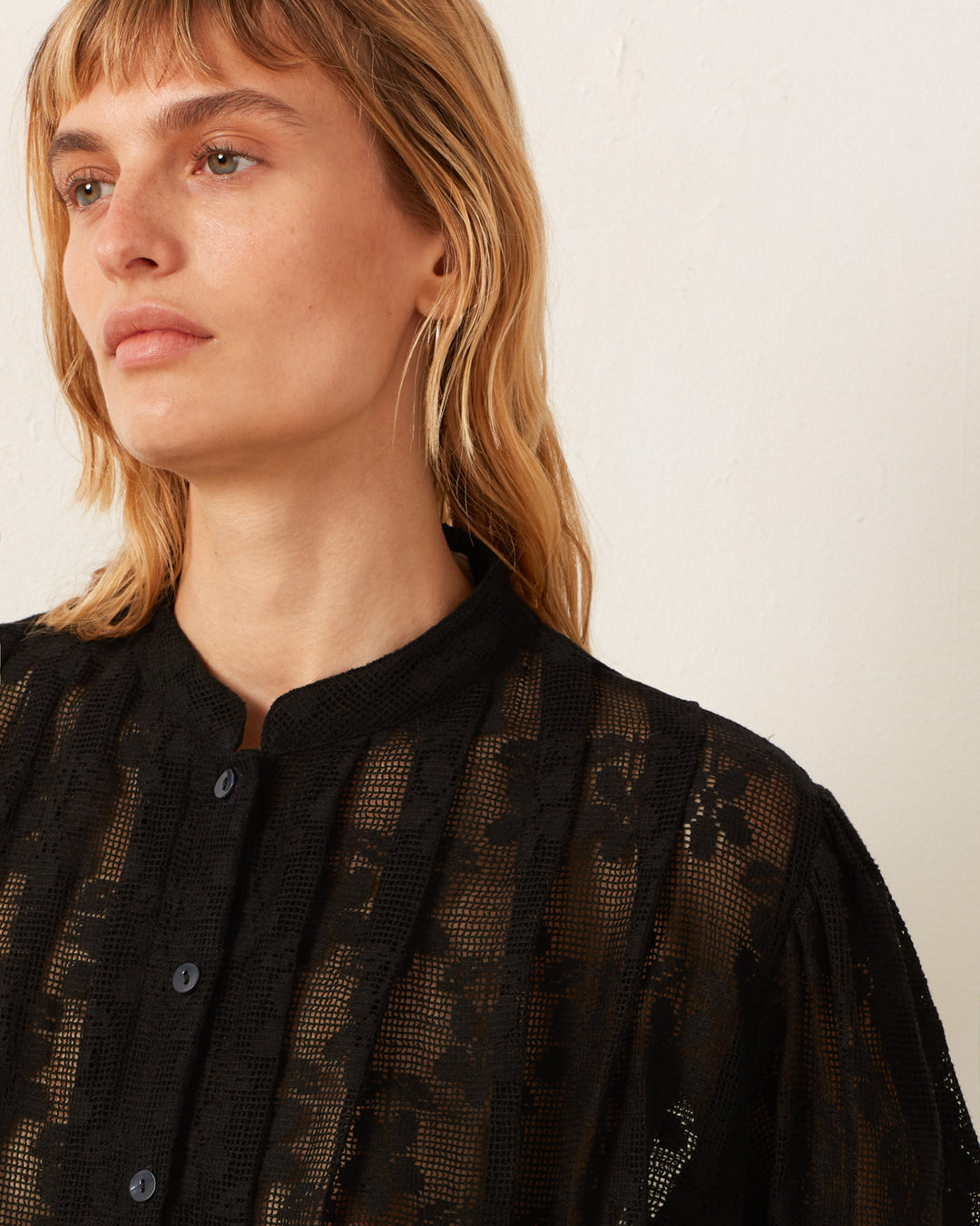 A person with shoulder-length blonde hair is wearing the Ryland Blackjack Daisy Shirt, a black cotton lace blouse featuring floral patterns that evoke a Victorian silhouette. They are looking slightly to the side against a light-colored background.