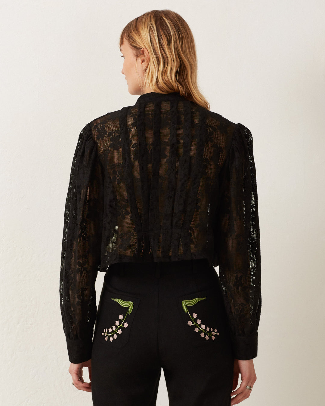 A person with medium-length hair is turned away from the camera, wearing the sheer black Ryland Blackjack Daisy Shirt and black pants featuring intricate floral embroidery on the back pockets. The background is a plain, light-colored wall.