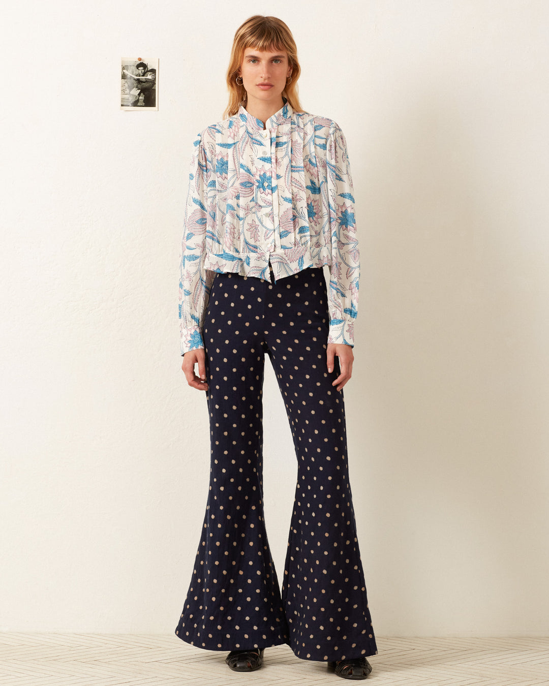 A person stands against a white wall, wearing the Ryland Blue Valentine Shirt and dark, hand-block printed bell-bottom pants. A small black-and-white photo is on the wall beside them.