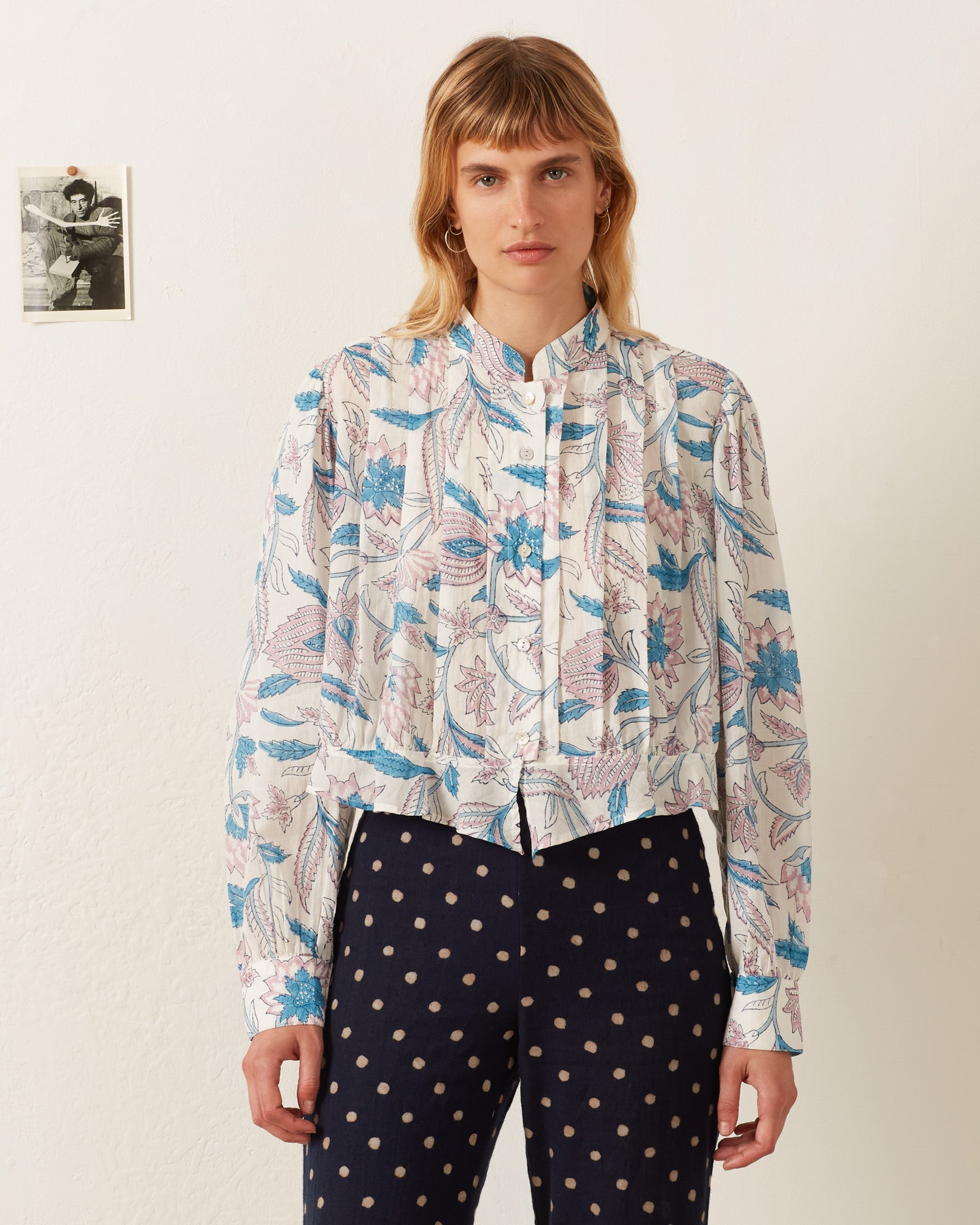 A person with shoulder-length hair wears the Ryland Blue Valentine Shirt paired with polka dot pants, showcasing a floral motif reminiscent of a Victorian silhouette, and stands in front of a white wall with a small black-and-white photo.