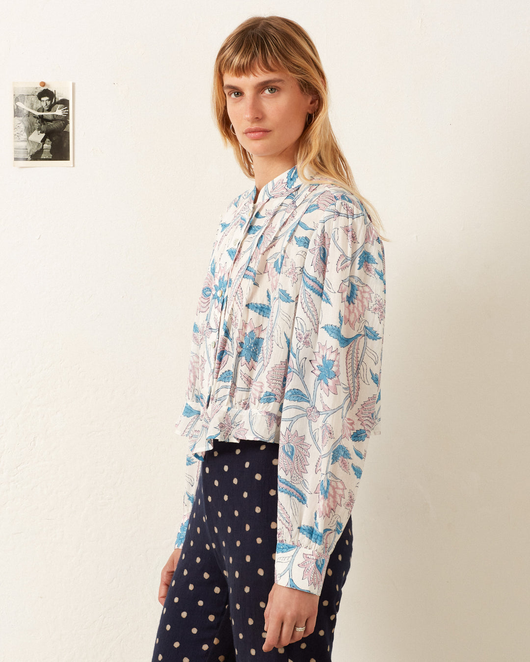 A person with long hair wearing the Ryland Blue Valentine Shirt, featuring a floral motif and long sleeves, and polka dot pants stands in a room where a small photo adorns the white wall.