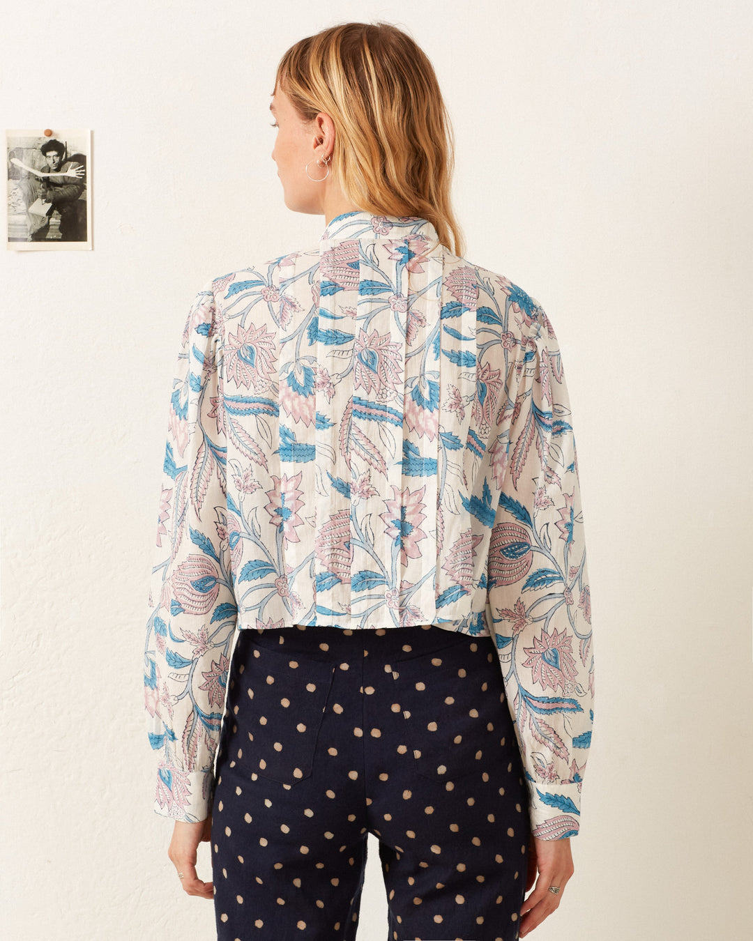 A person stands facing a wall, wearing the Ryland Blue Valentine Shirt and polka dot pants. A small black-and-white photograph is pinned to the wall, evoking the delicate charm of a Victorian silhouette.