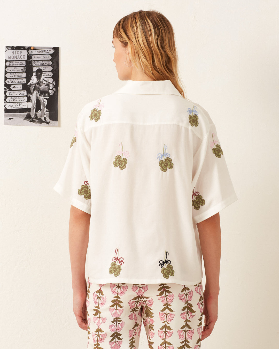 A person with shoulder-length hair stands with their back to the camera, wearing the Stevie Lucky Clover Shirt — a white hand-embroidered top adorned with floral designs — paired with patterned pants in a room featuring a white wall and a black-and-white poster.