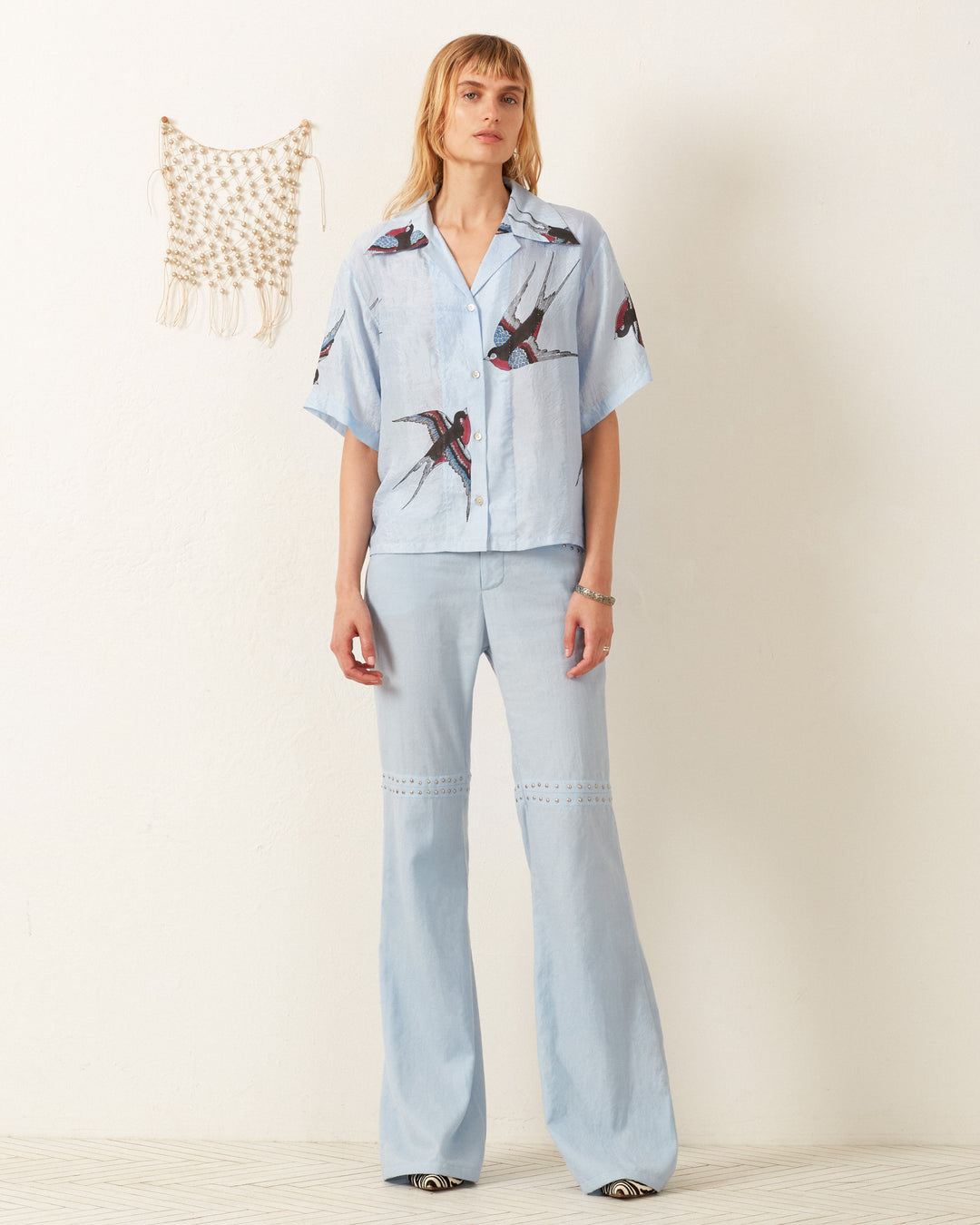 Against a plain white wall, a person wears the Stevie Swallow Silk Shirt, featuring a light blue hand-block printed design with swallow prints paired with wide-legged pants. A small macrame wall hanging is visible on the left above the wooden floor.