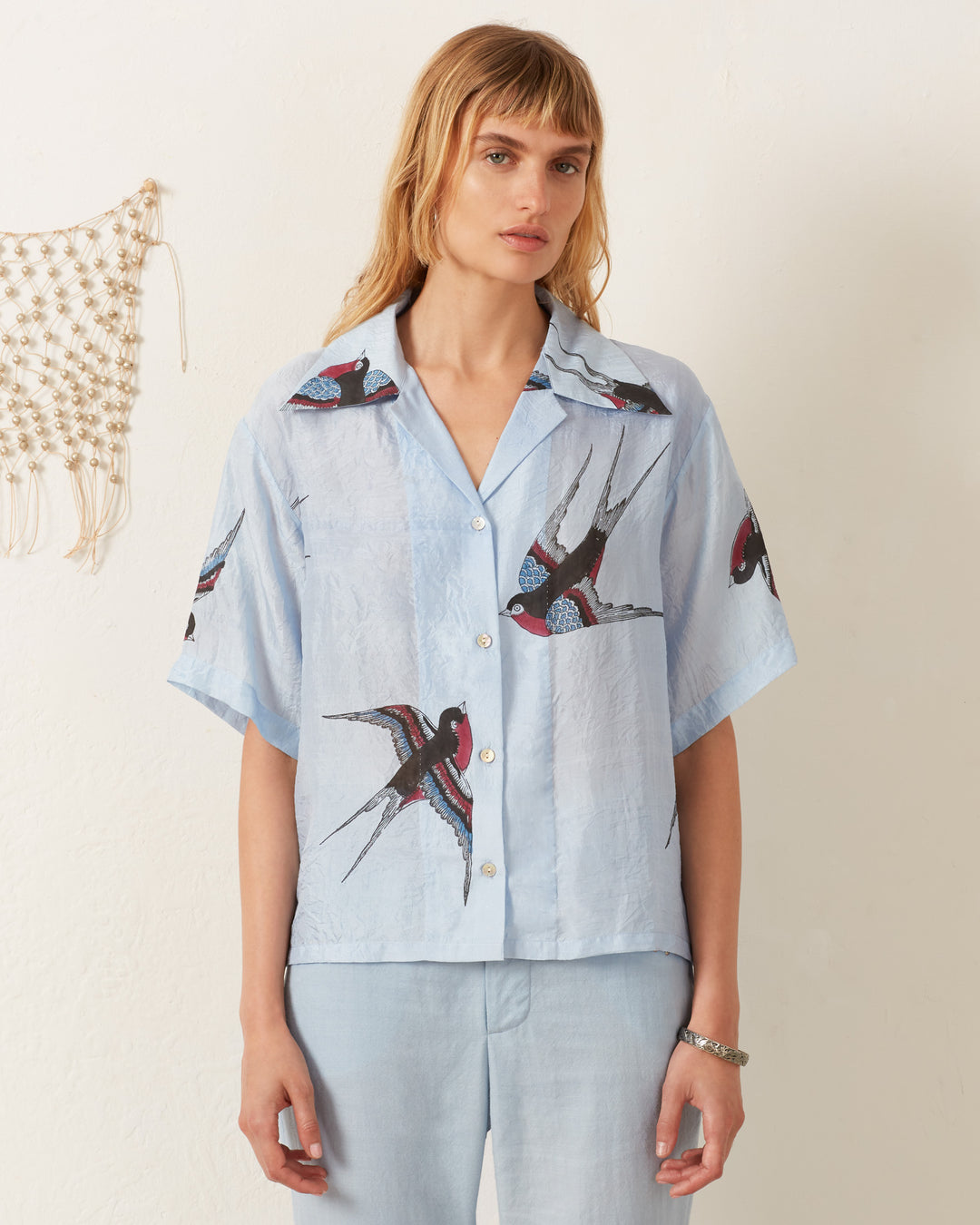 A person with long hair wears the Stevie Swallow Silk Shirt, a light blue Hawaiian-style shirt featuring a swallow bird pattern. They stand against an off-white wall with a small macramé decoration hanging nearby.