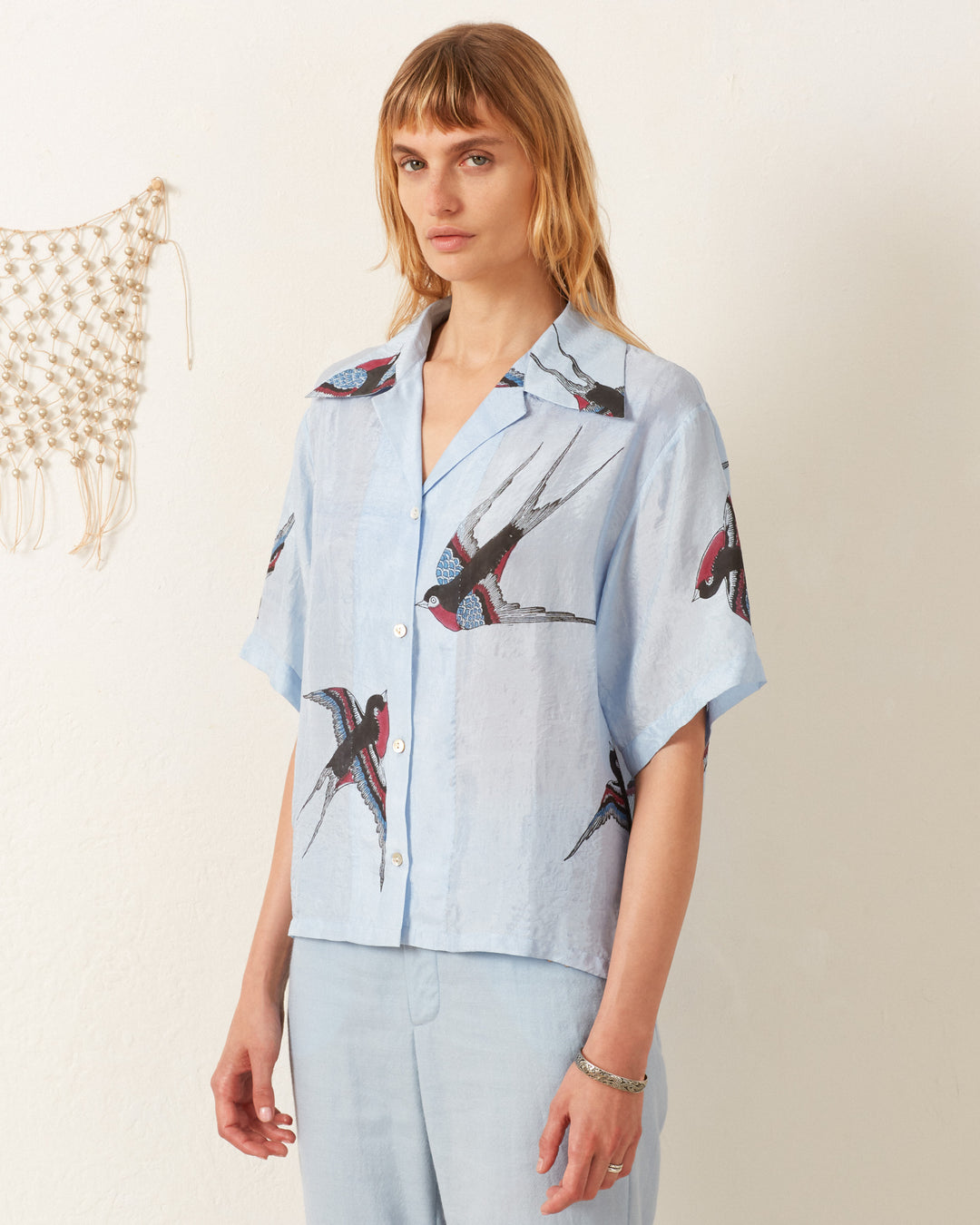 A person with long hair wearing a vintage Stevie Swallow Silk Shirt featuring bird prints and light blue pants stands in front of a white wall adorned with a macrame piece.