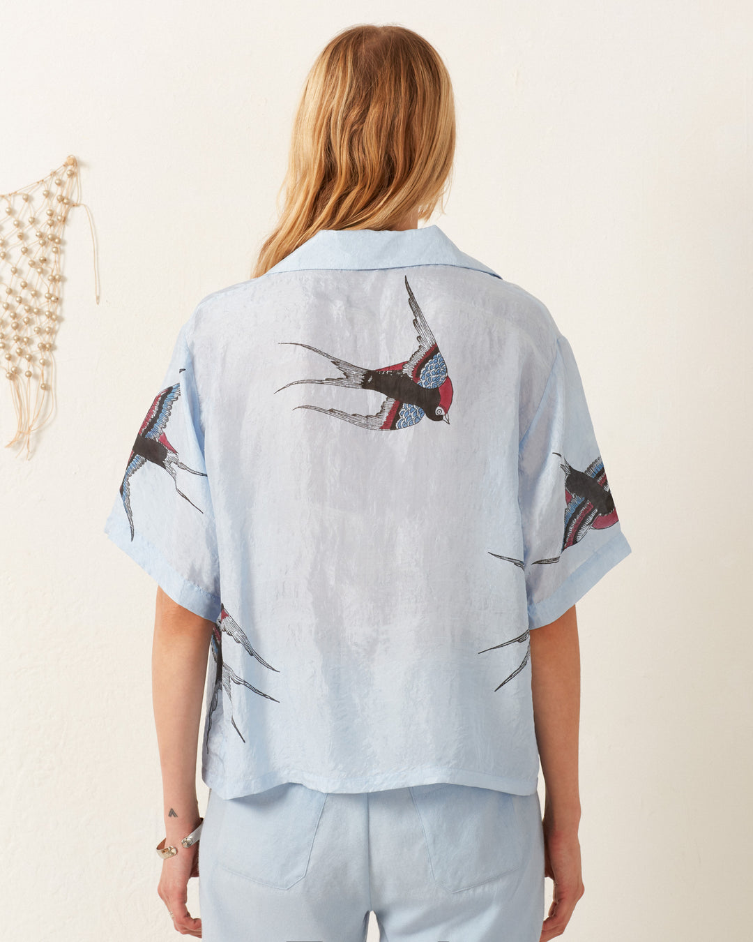 A person with long blond hair faces away, wearing the Stevie Swallow Silk Shirt in light blue adorned with swallow illustrations, paired with matching pants. A macramé wall hanging is partially visible on the left.