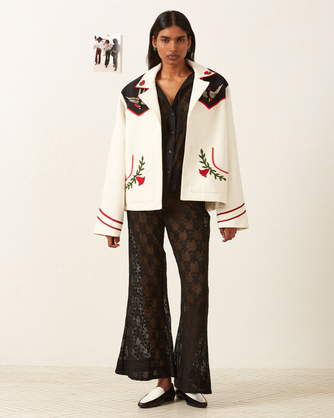 A person stands against a white wall wearing the Western Royal Flush Jacket, featuring hand-embroidered designs with black and red detailing, over a black lace outfit. They have long dark hair and strike a confident pose, while a small picture is visible on the wall behind them.