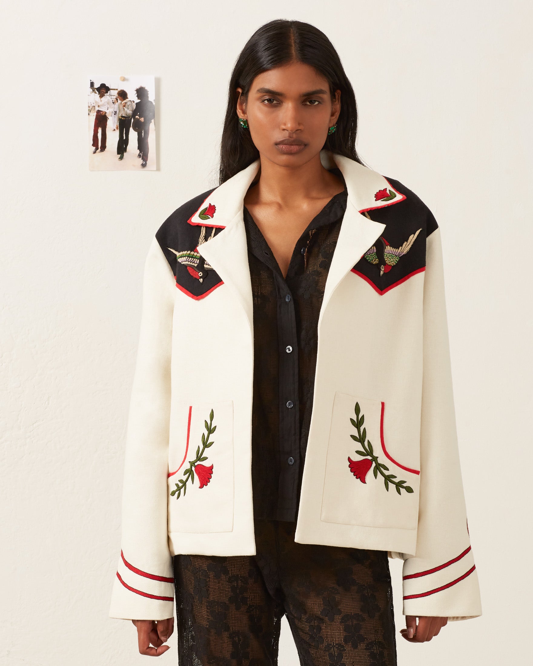 A person with long dark hair wears the Western Royal Flush Jacket, a white garment with red and green floral embroidery, over a black lace outfit. The jacket features large lapels and intricate hand-embroidered decorative patterns. A small photo is pinned to the wall behind them.