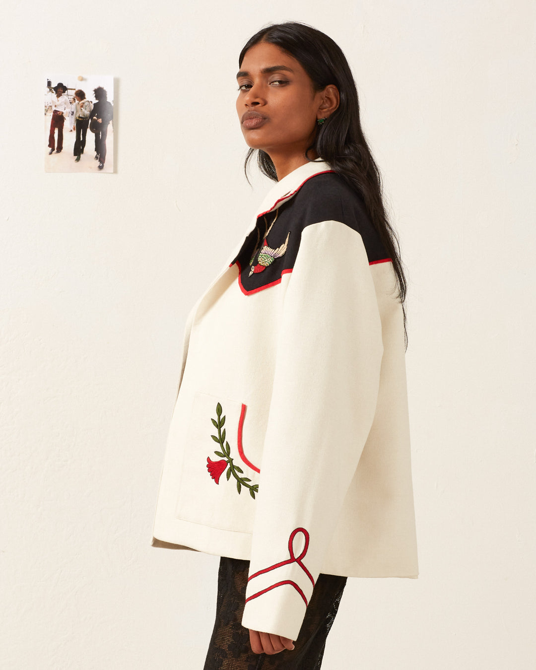 A person with long dark hair stands in profile, sporting the Western Royal Flush Jacket, a chic silk piece featuring black and red accents with Western-inspired floral embroidery. A small photo is pinned to the wall in the background.