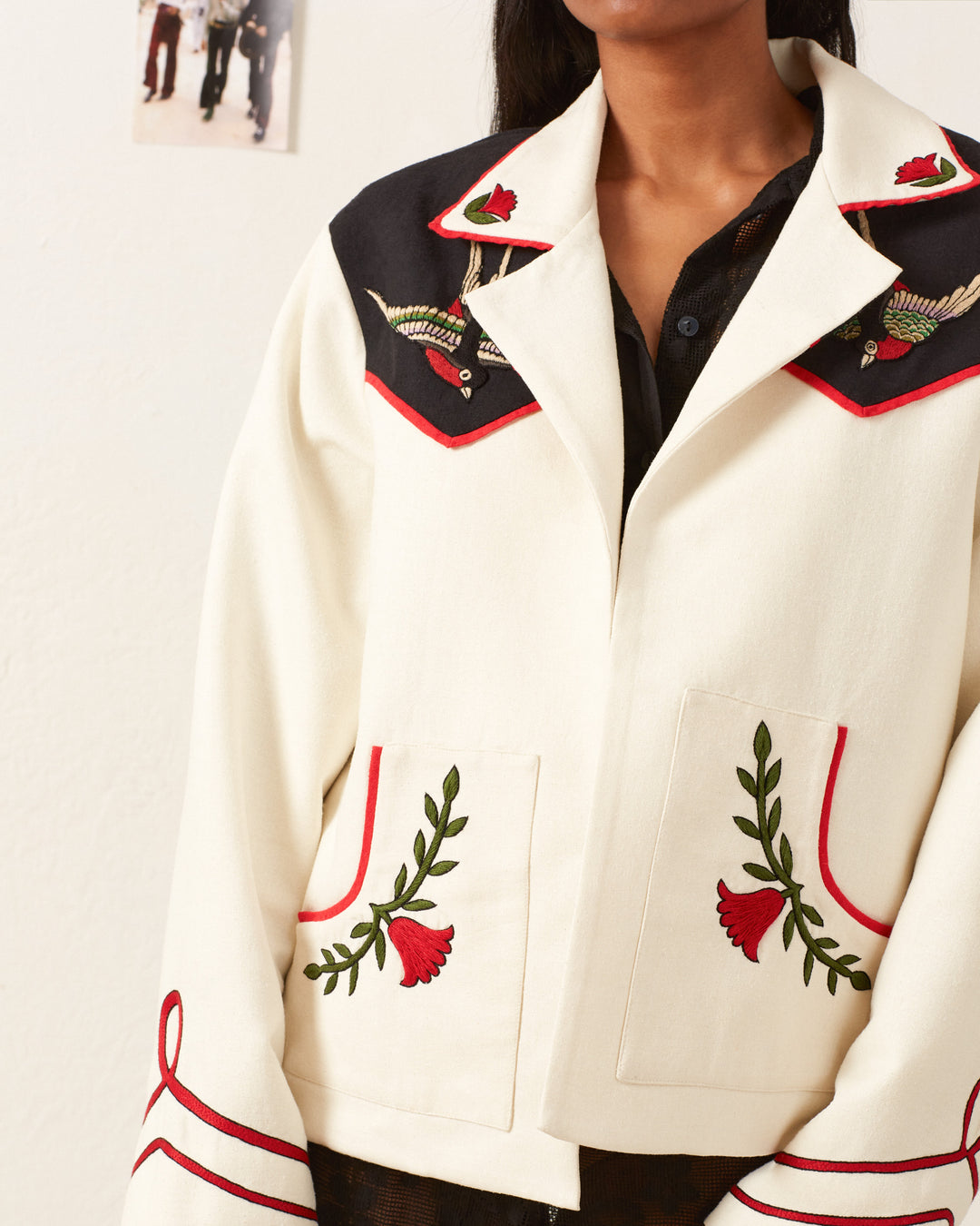 A person wearing the Western Royal Flush Jacket, a Western-inspired white jacket adorned with red and green floral embroidery and black accents. This hand-embroidered silk jacket boasts two large front pockets and intricate patterns on the shoulders. A small photo is pinned to the wall behind them.