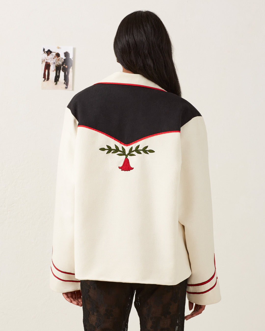 Rear view of a person with long hair wearing the Western Royal Flush Jacket, cream-colored with black and red accents. It features a hand-embroidered red rose with green leaves on the back. A small photo is pinned to a white wall in the background.