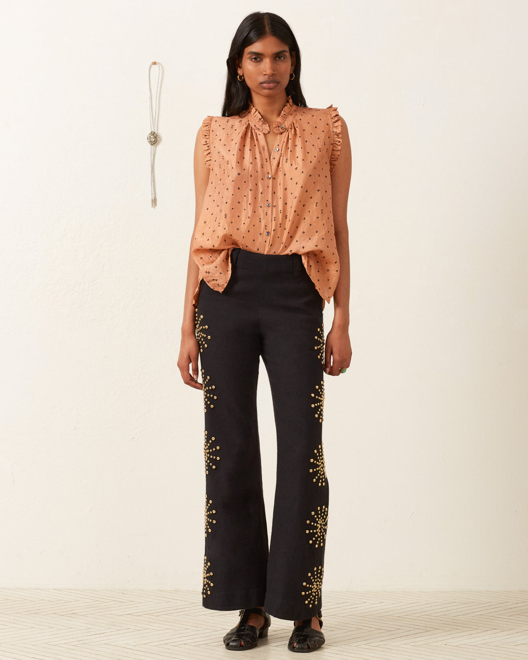 Against a plain white background, an individual wears the Wylder Ace Silk Shirt with polka dots and black flared pants featuring hand-printed floral designs. A necklace hangs on the wall beside them.