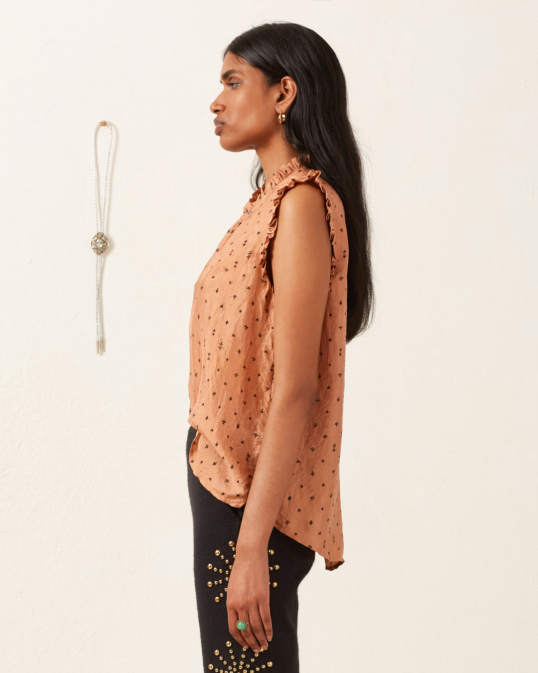 A person with long, dark hair stands in profile against a plain background, wearing a rust-colored Wylder Ace Silk Shirt with ruffled edges and black pants adorned with small gold embellishments. A decorative necklace hangs on the wall.