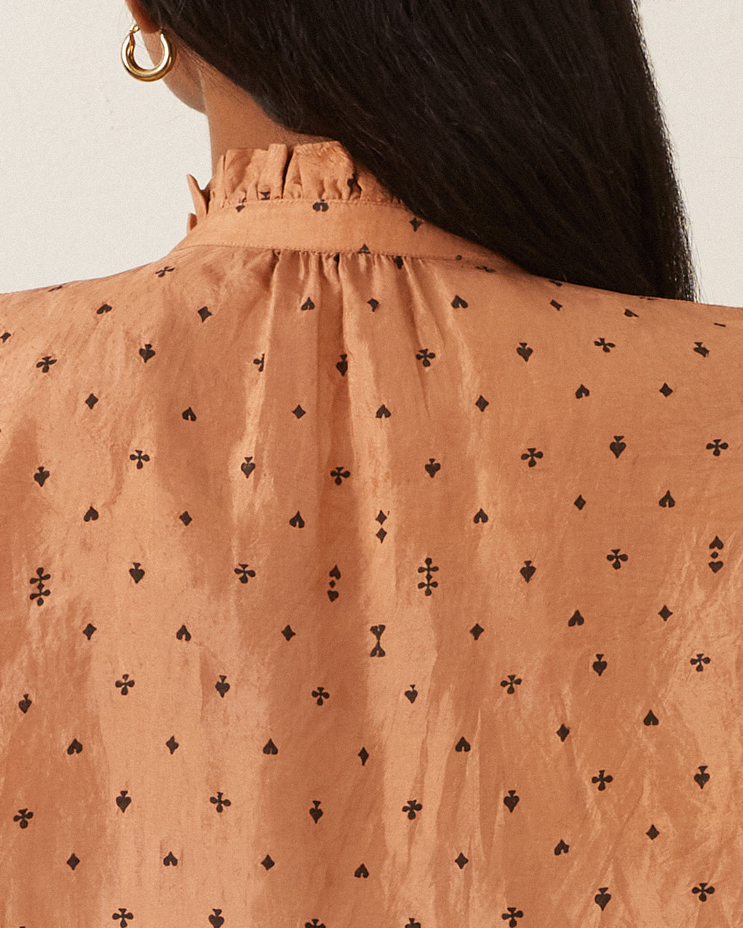 A woman with long dark hair is wearing the Wylder Ace Silk Shirt, a tan blouse with a high ruffled collar and hand-printed black diamond and cross patterns. Her gold hoop earrings gleam as she turns away from the camera, highlighting the blouse's elegant texture that evokes the feel of hand-loomed silk.