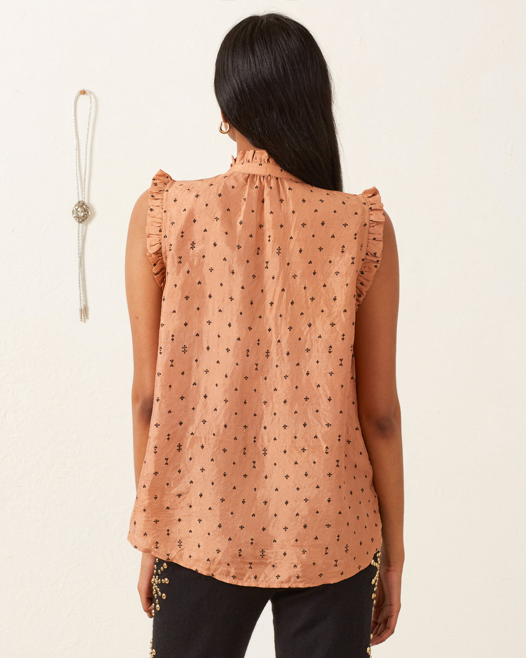 A person with long black hair is wearing the Wylder Ace Silk Shirt, a hand-loomed sleeveless peach blouse featuring ruffled edges and small black patterns, paired with black pants. They are standing against a white wall, with a beaded necklace hanging on the left side.