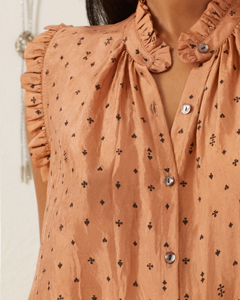 A close-up of a person wearing the Wylder Ace Silk Shirt, which is peach-colored and features hand-printed small black shapes. This sleeveless blouse includes ruffled collar edges, button-down detailing, and a soft, textured fabric that adds an elegant touch.