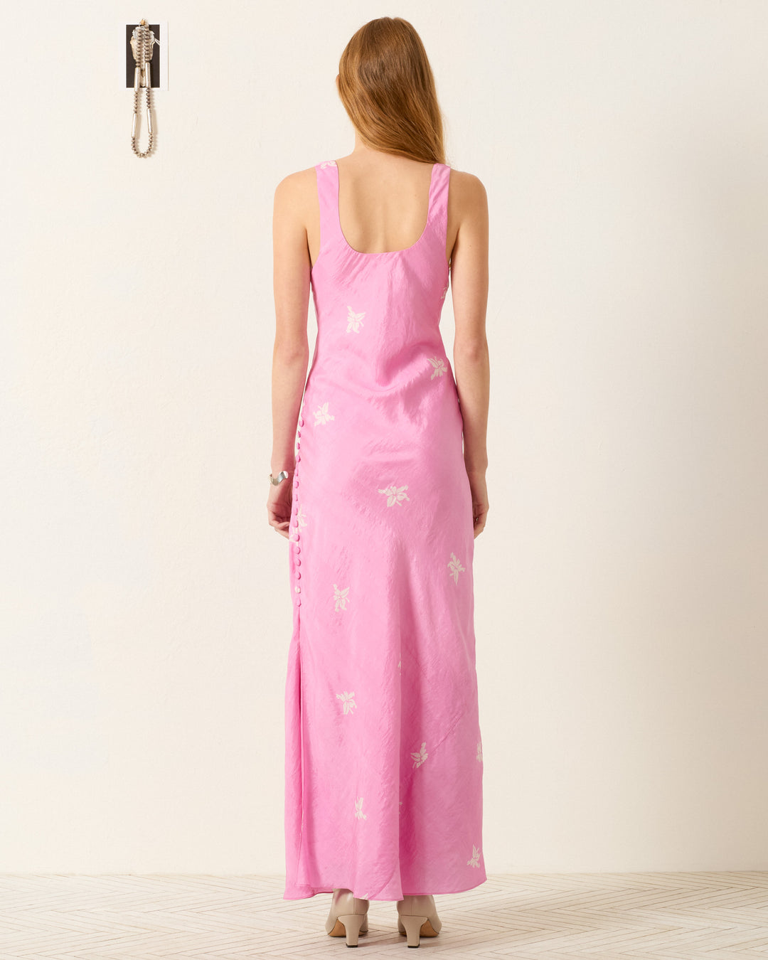 A person with long hair faces away, wearing the Adele Pink Jasmine Dress—a sleeveless, hand-block-printed, floor-length pink dress with a white floral motif and beige shoes. A wall-mounted phone appears in the upper left corner.