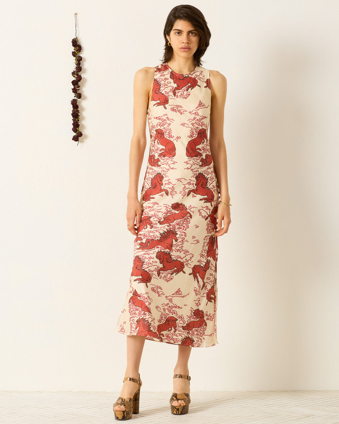 A woman wears the Adelaide Rouge Pegasus Dress—an ankle-length, sleeveless, hand-loomed silk dress with a red and beige animal print—while standing by a white wall next to a hanging decoration.