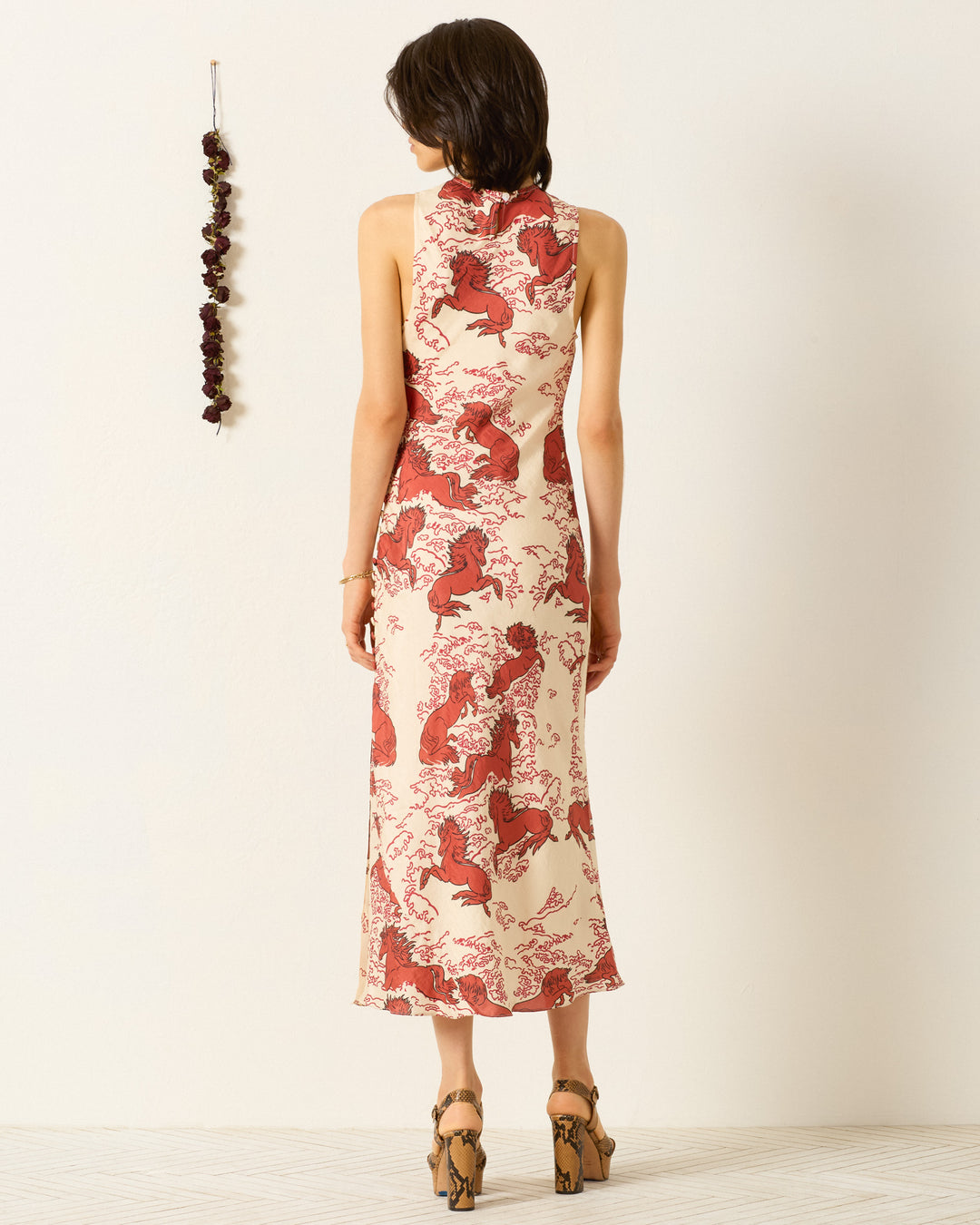 A person faces away wearing the Adelaide Rouge Pegasus Dress, a bias-cut slip dress featuring a red and cream mythical horse motif, paired with wooden platform heels. Dried flowers are displayed on the nearby wall.