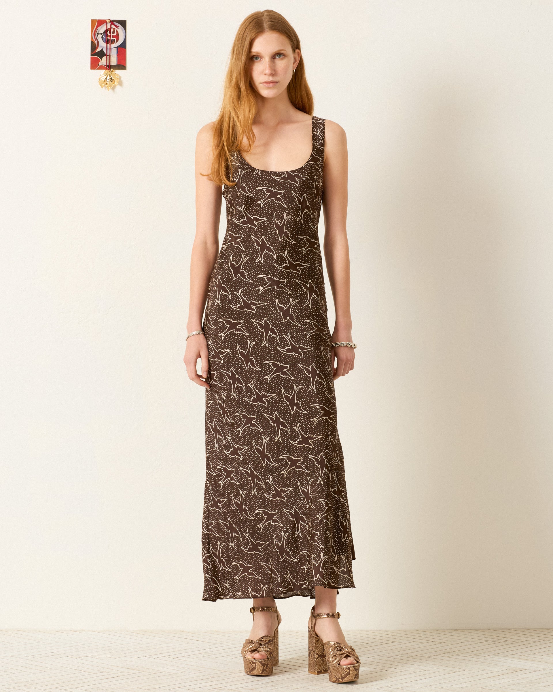 A woman stands against a plain wall, wearing the Adele Clove Swallow Silk Dress with a bird pattern and platform sandals.