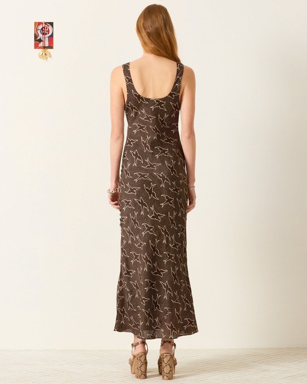 A woman faces away, wearing the Adele Clove Swallow Silk Dress—a sleeveless, ankle-length artisan piece in brown with a white abstract pattern—paired with tan platform heels.