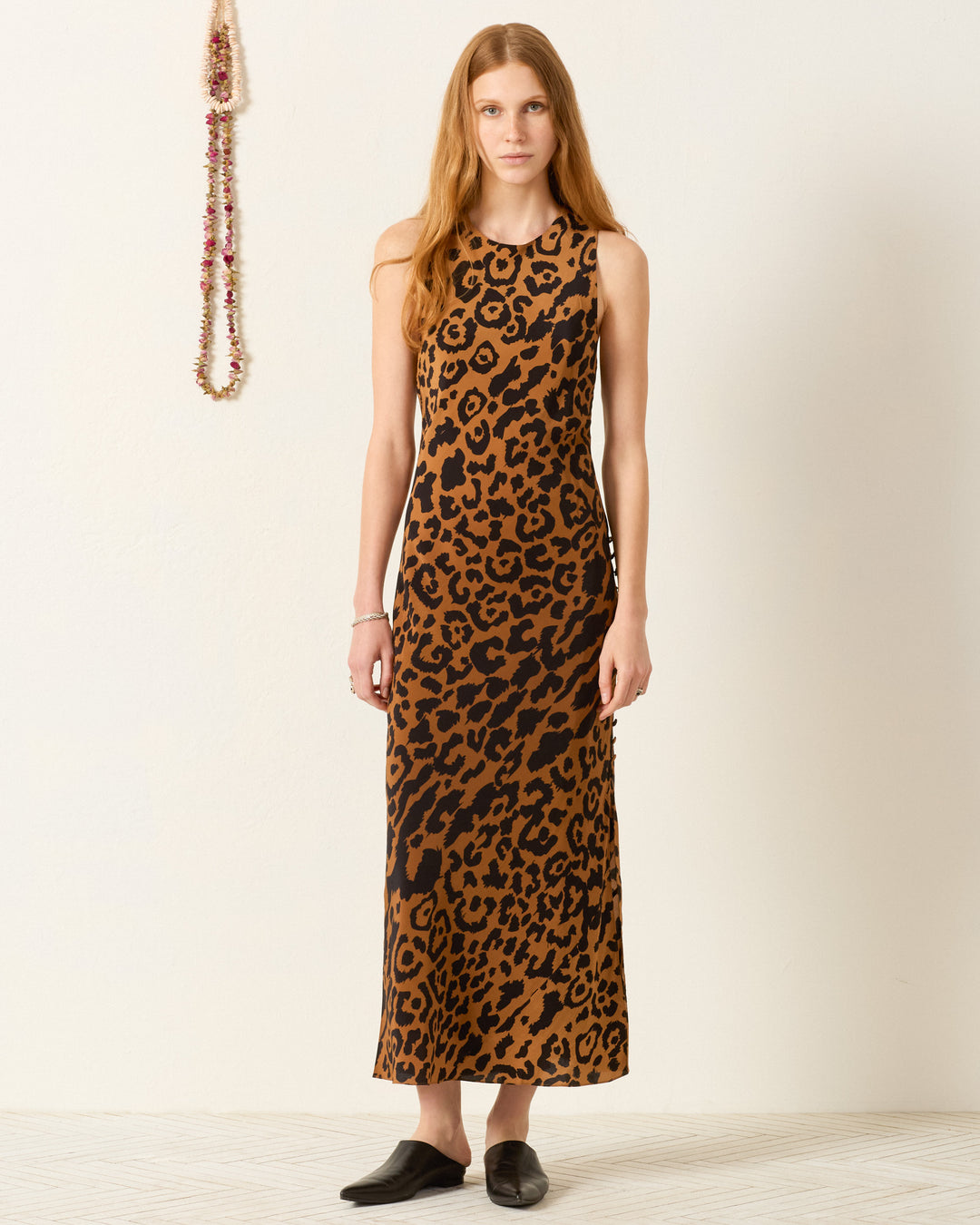 A woman stands indoors in the Adelaide Leopard Silk Dress, a sleek, bias-cut slip dress with a leopard print, paired with black slip-on shoes. A beaded necklace decorates the wall behind her.
