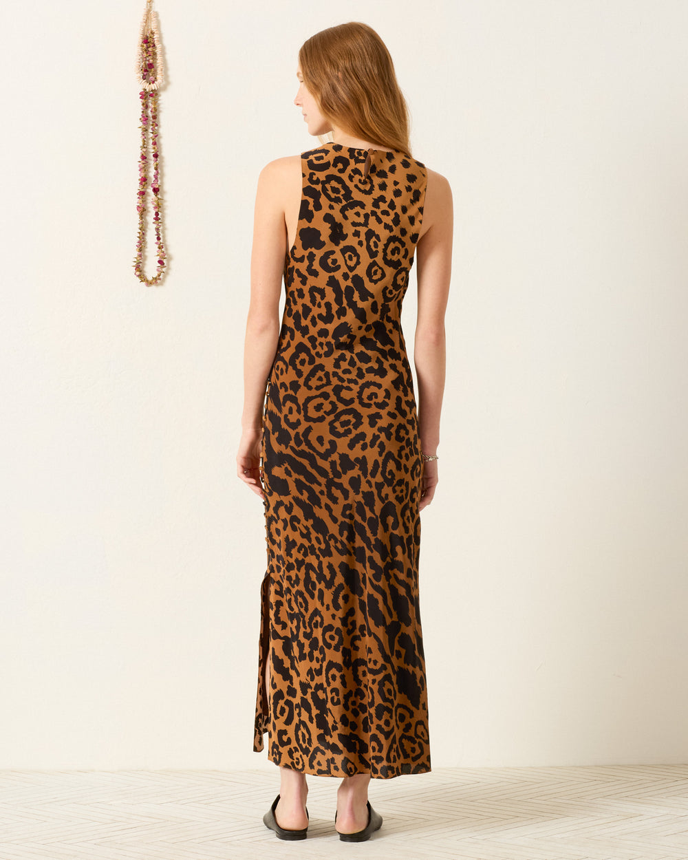 A woman stands facing away, wearing the Adelaide Leopard Silk Dress, an ankle-length bias-cut slip dress in leopard print with black sandals. A beaded necklace is displayed on the wall beside her.