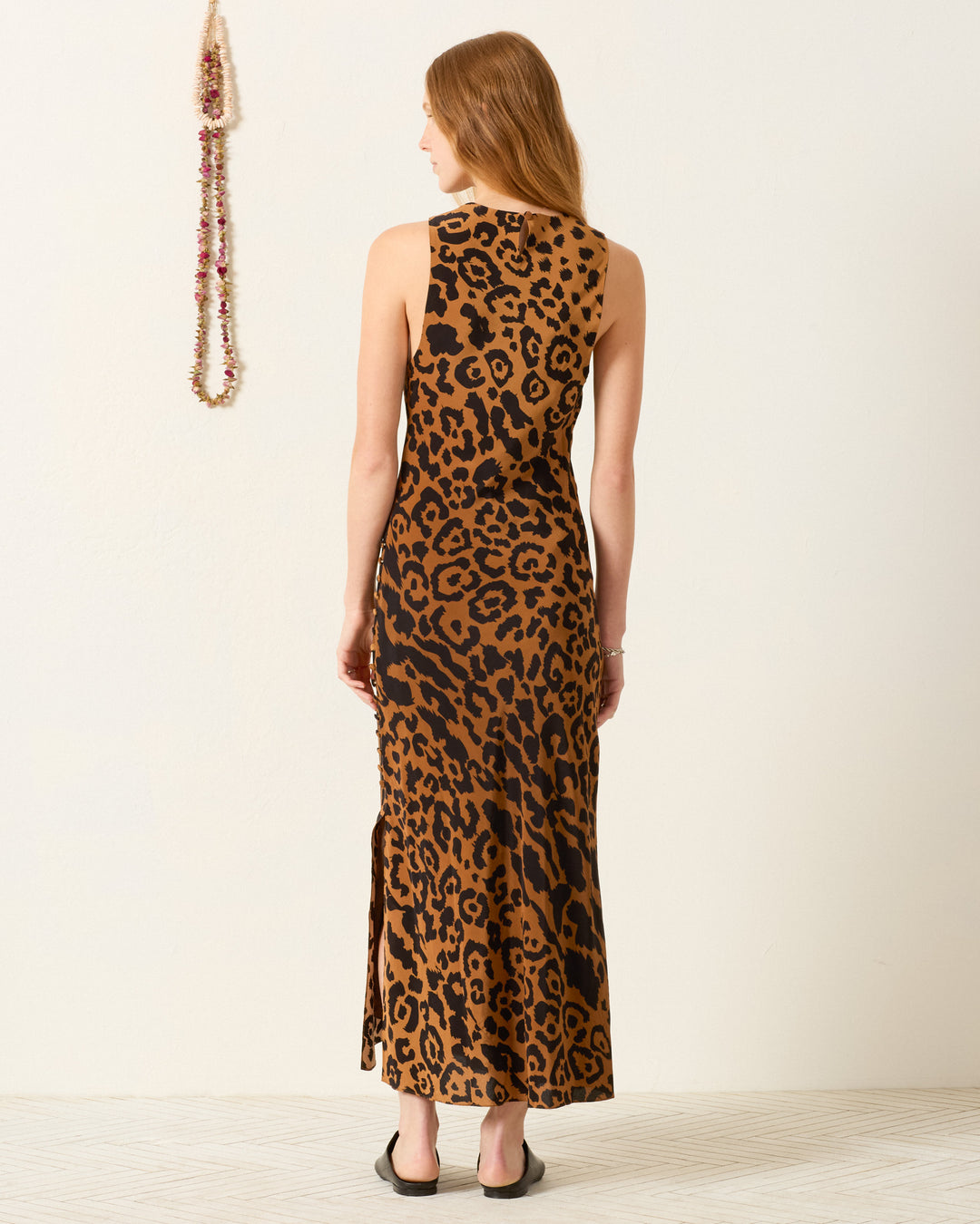 A woman stands facing away, wearing the Adelaide Leopard Silk Dress, an ankle-length bias-cut slip dress in leopard print with black sandals. A beaded necklace is displayed on the wall beside her.