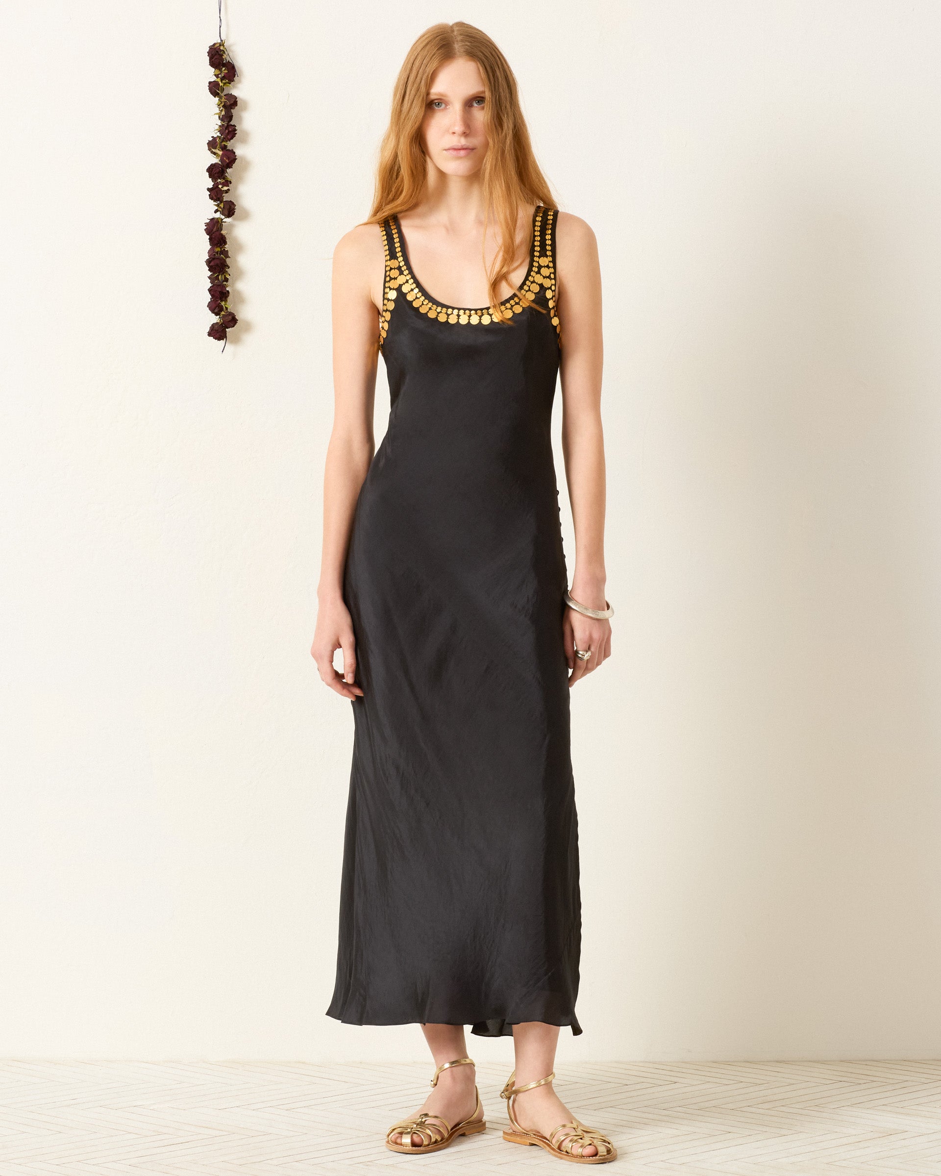 A woman stands against a white wall wearing the Adele Gilded Coin Dress, an artisan-made long black sleeveless dress with gold neckline detailing, paired with gold sandals. Dried flowers hang on the wall beside her.