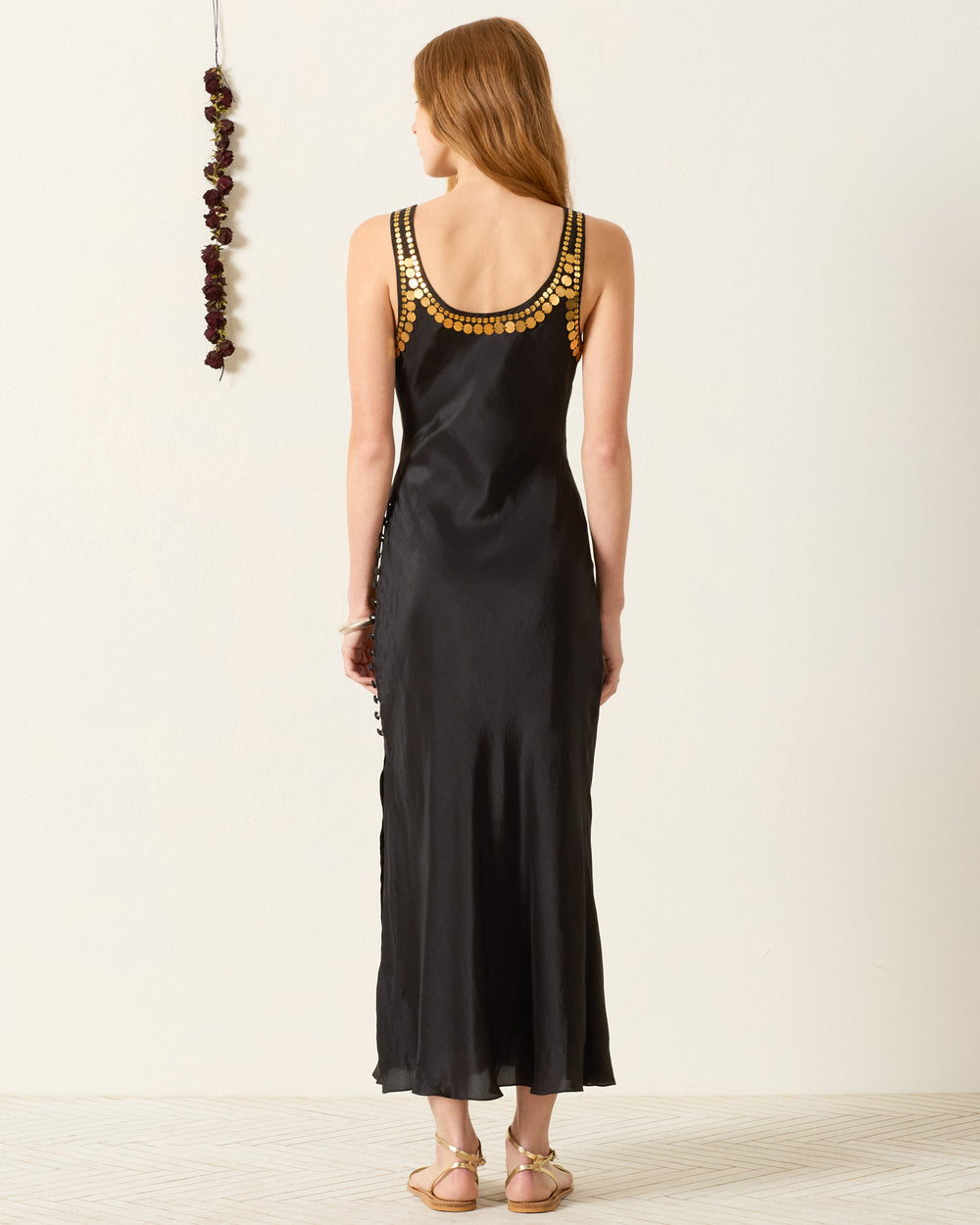 A woman faces away, wearing the artisan-made Adele Gilded Coin Dress—an ankle-length black dress with gold shoulder embellishments—and flat sandals. A string of dried flowers hangs on the wall.