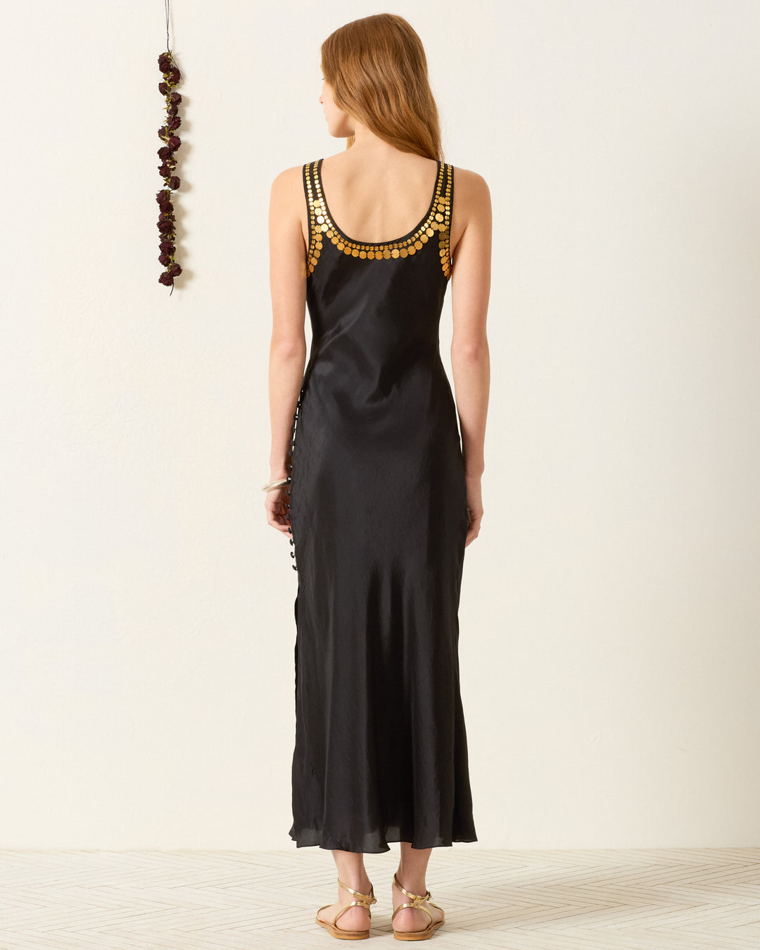 A woman faces away, wearing the artisan-made Adele Gilded Coin Dress—an ankle-length black dress with gold shoulder embellishments—and flat sandals. A string of dried flowers hangs on the wall.