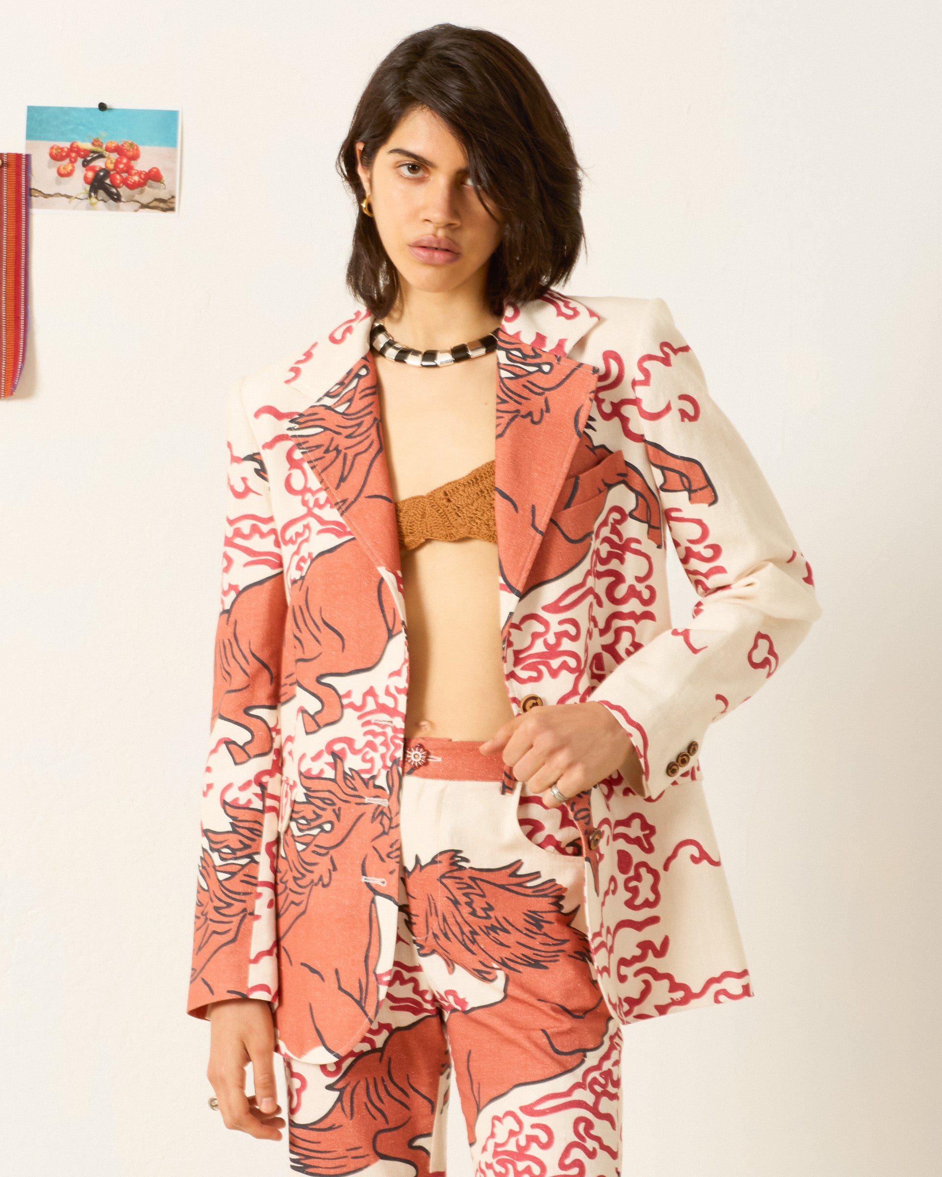 A woman stands against a white wall wearing the Afternoon Rouge Pegasus Blazer, a brown bralette, and a black beaded necklace.