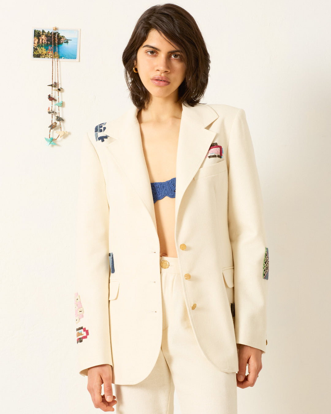 A woman wearing the Afternoon Abstract Tile Blazer, a cream-colored hand-beaded jacket with embroidered patches, stands against a white wall. A blue bralette peeks from under her blazer with small wall decorations behind her.