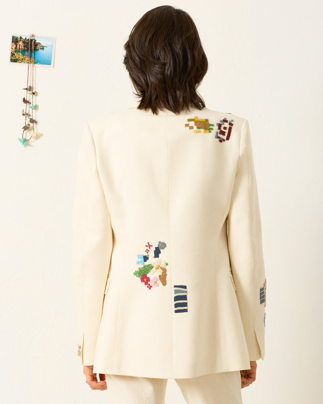 A person wearing the Afternoon Abstract Tile Blazer, a cream blazer with colorful beaded patterns, stands with their back to the camera; wall decor appears on the left.