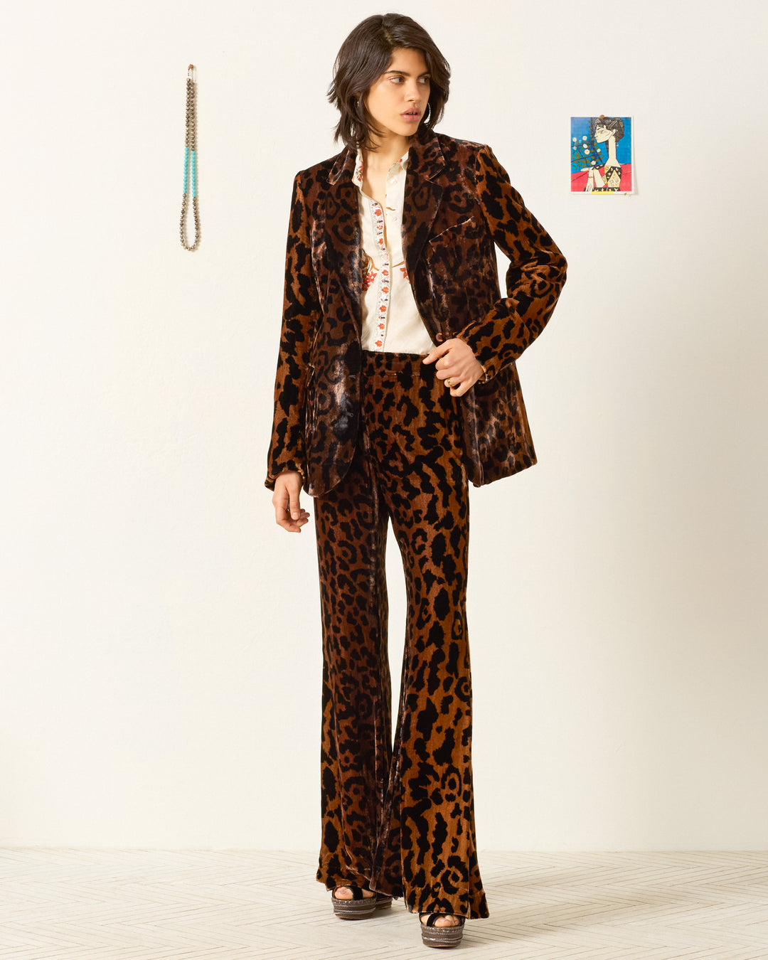 Indoors, a person wears the Afternoon Leopard Blazer with flared pants and a white shirt. The blazer's notched lapel adds classic style to the bold leopard print. They stand near a wall decorated with two small accents.