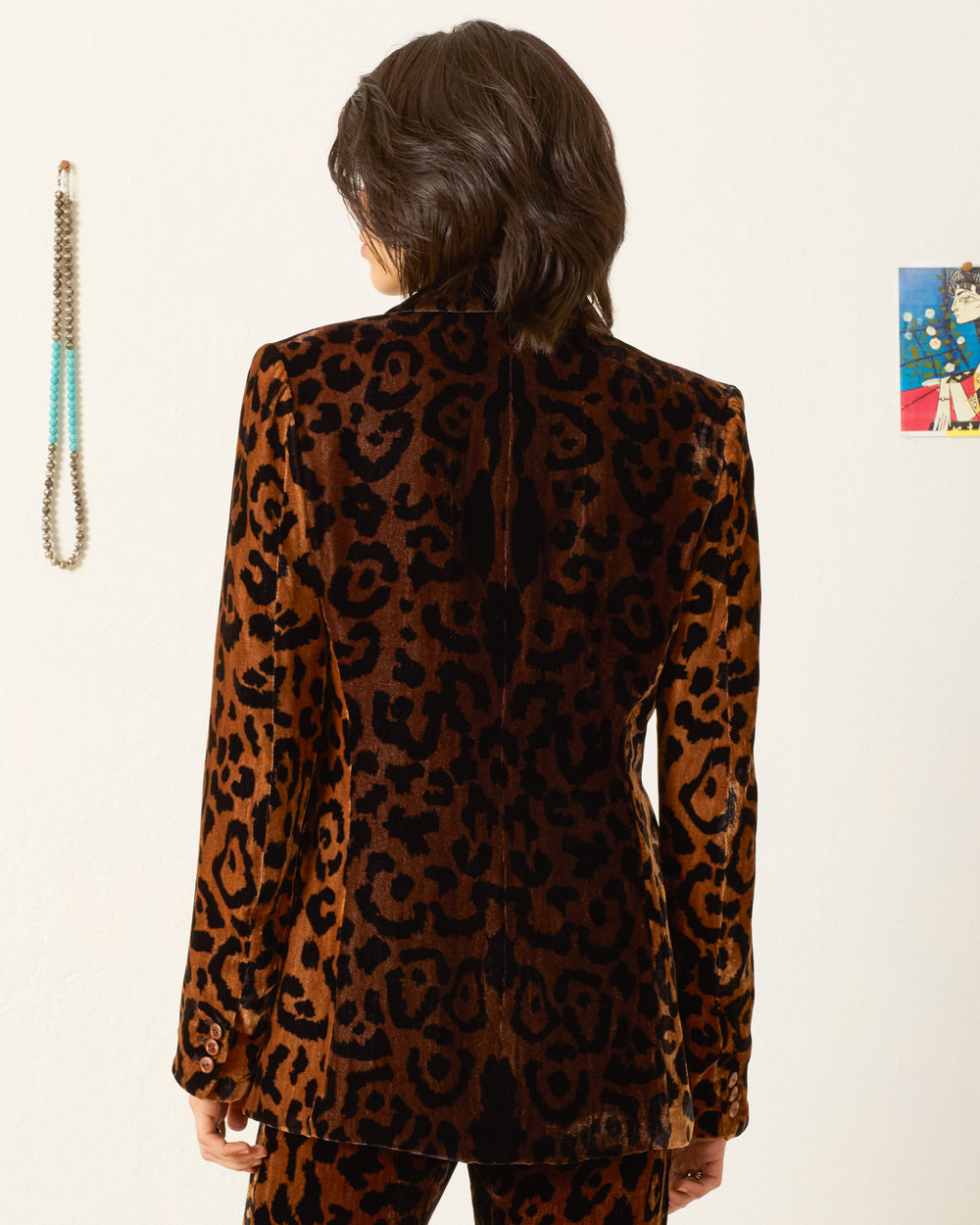 A person with shoulder-length dark hair stands facing away, wearing the Afternoon Leopard Blazer—a velvet blazer with a notched lapel and matching pants. A beaded necklace and a small colorful picture decorate the wall.