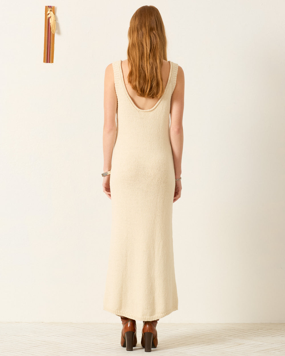 A person with long, light brown hair stands facing away, wearing the Amery Lovebird Knit Dress and brown heeled shoes against a plain, light background.
