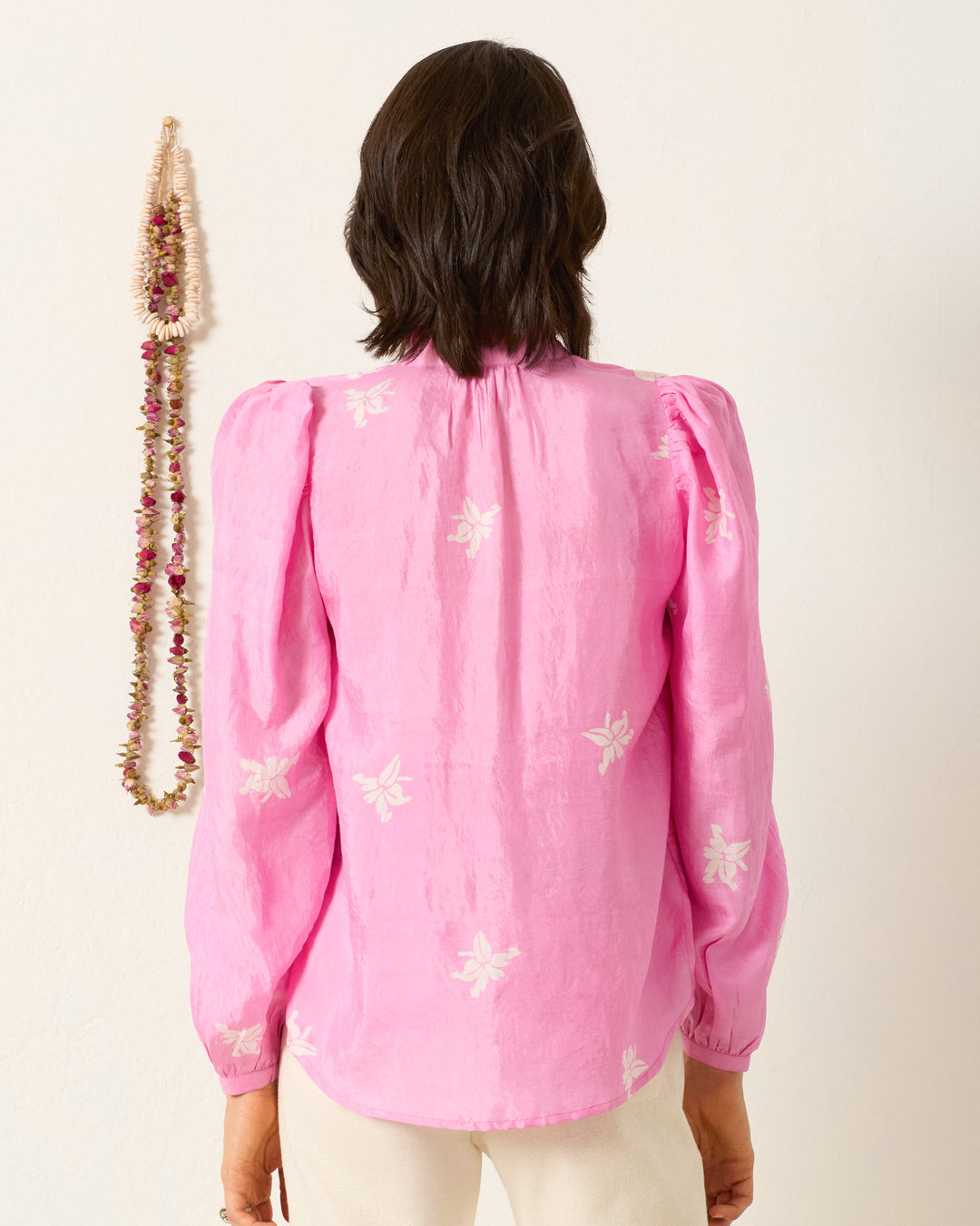 Someone wearing the Annabel Pink Jasmine Shirt, featuring white floral motifs, stands facing a wall decorated with a beaded necklace.
