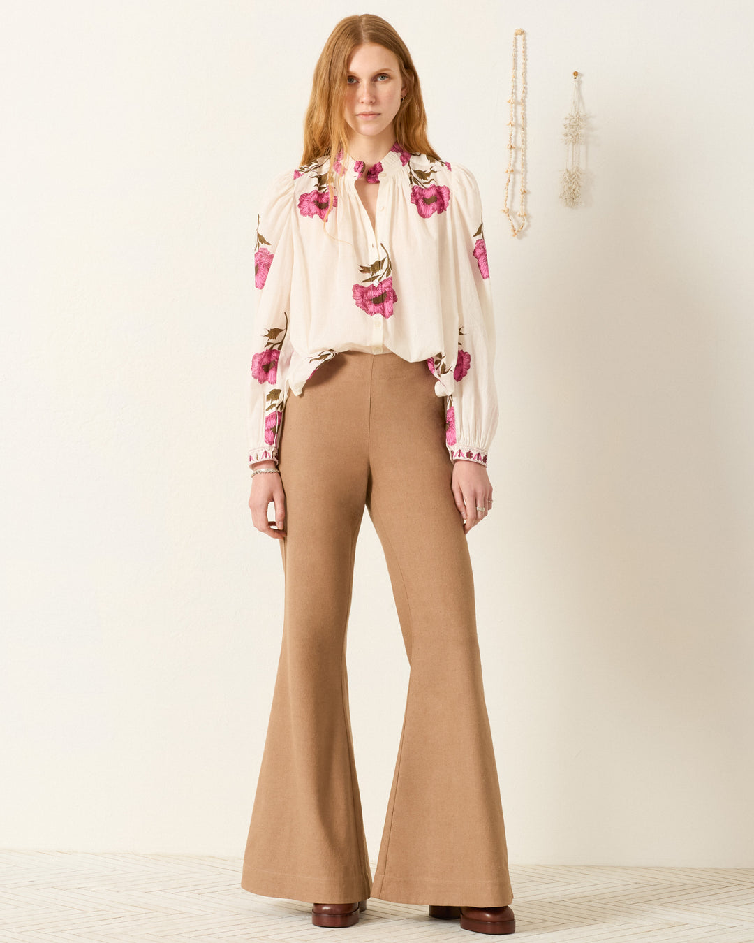 A woman stands indoors wearing the Annabel Fuchsia Poppy Shirt—a white hand-block-printed cotton voile shirt with pink floral designs—paired with high-waisted tan flared pants and brown platform shoes. Neutral wall decor in background.