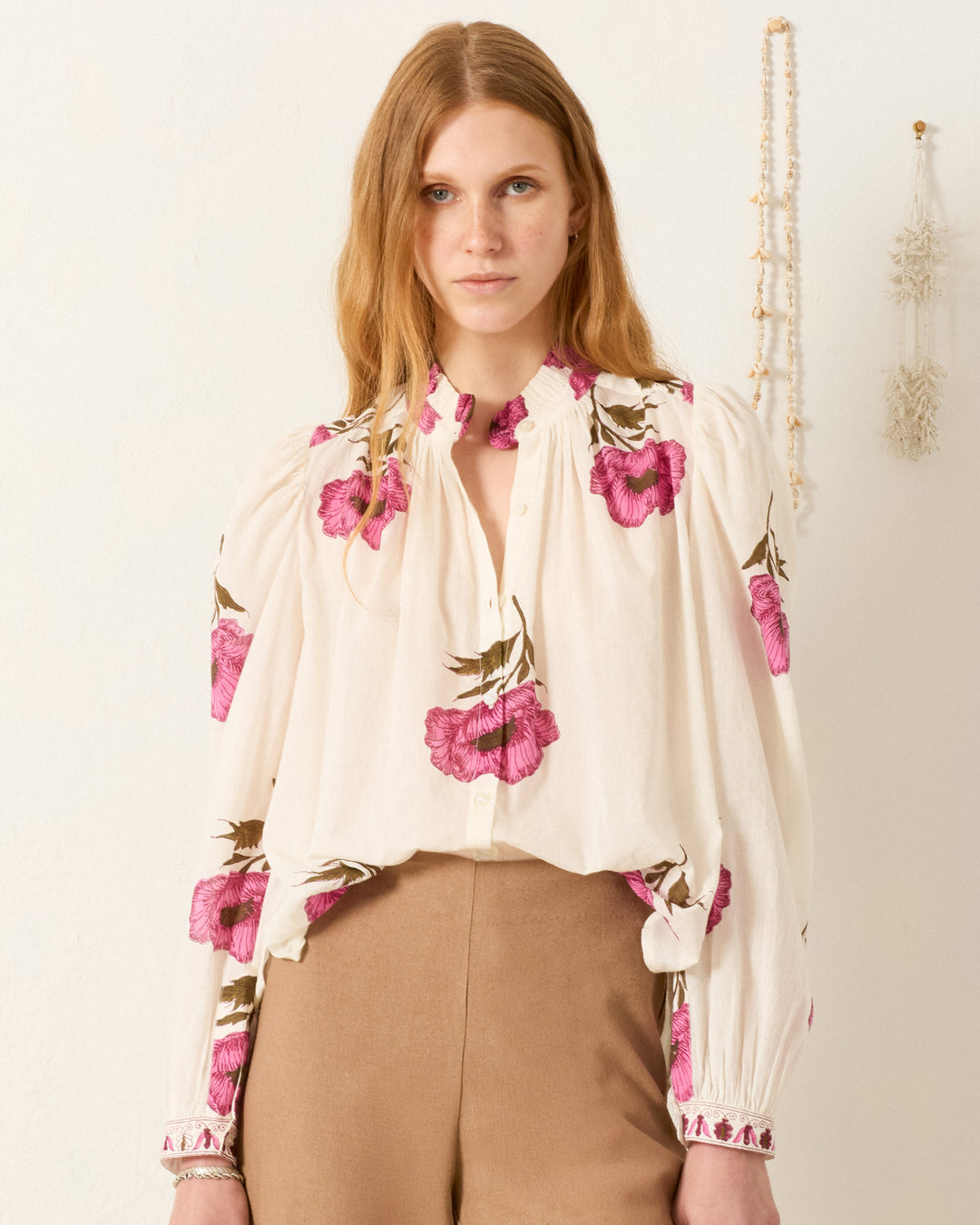 A woman with long, light brown hair wears the cream Annabel Fuchsia Poppy Shirt, featuring large pink floral prints and a ruffled collar, paired with tan high-waisted trousers, standing against a light-colored wall with hanging decor.