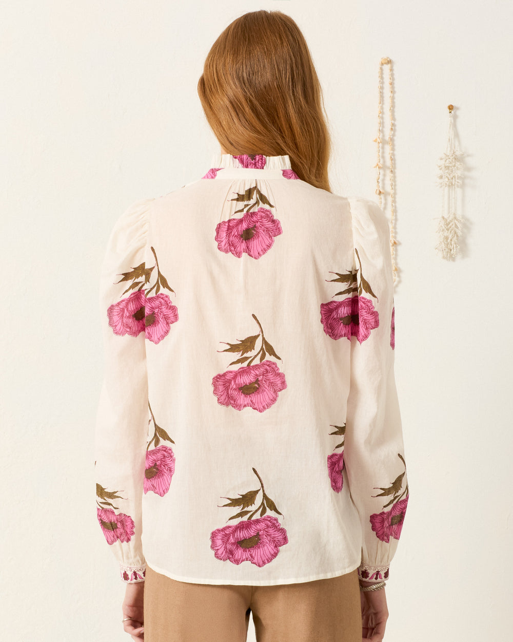 A person with long, light brown hair is seen from the back, wearing the Annabel Fuchsia Poppy Shirt—a hand-block-printed cotton voile shirt featuring pink floral patterns and a delicate ruffled collar.