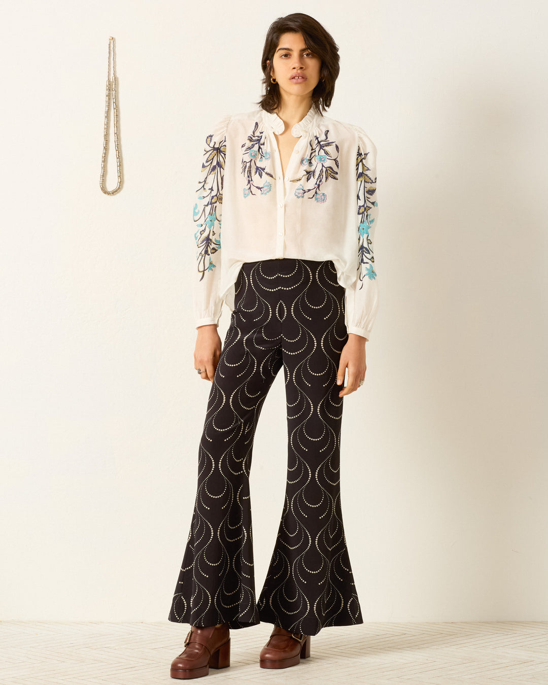 Wearing the Annabel Misted Lily Shirt—a white cotton-silk blend with floral embroidery on the sleeves—paired with black dotted flared pants and brown platform shoes, a person stands against a plain wall.
