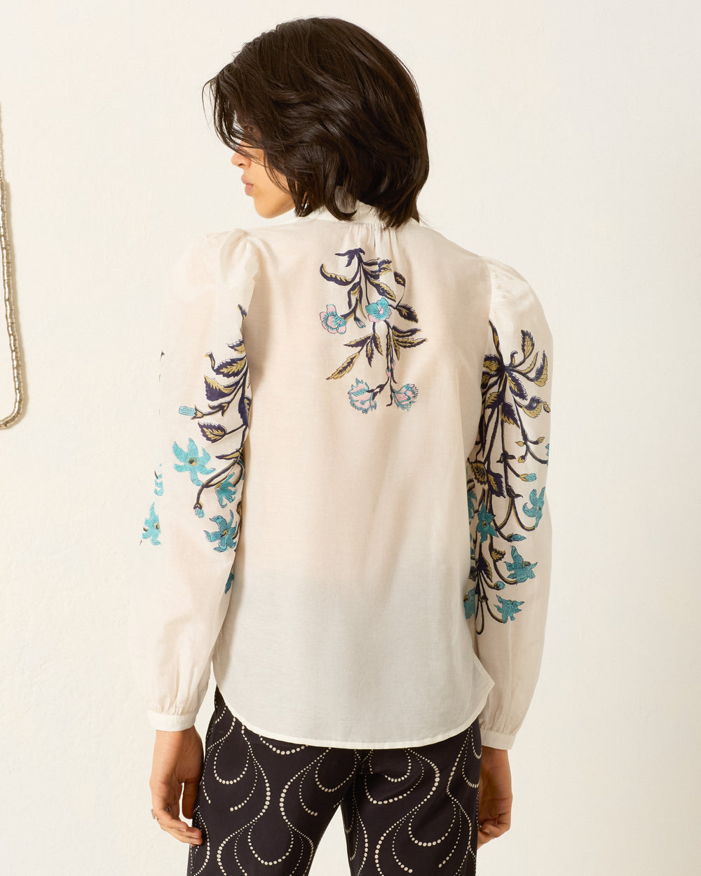 A person stands facing away, wearing the Annabel Misted Lily Shirt—a sheer cotton-silk blend with blue floral embroidery on the back and sleeves—paired with black pants featuring a white wavy pattern.