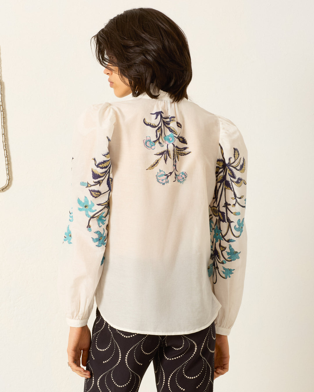 A person stands facing away, wearing the Annabel Misted Lily Shirt—a sheer cotton-silk blend with blue floral embroidery on the back and sleeves—paired with black pants featuring a white wavy pattern.